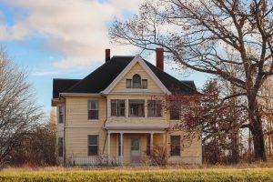 Photograph of an old yellow house, to the right of which stands a tall tree with long branches. The leaves are an Autumnal orange, and many have fallen to the ground.